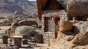 A rustic stone building with a thatched roof, large rocks surrounding the area, and a patio with chairs. - Canyon Lodge (Fish River Canyon)