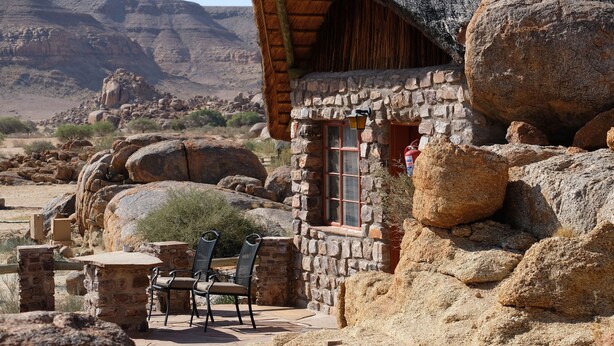 A rustic stone building with a thatched roof, large rocks surrounding the area, and a patio with chairs. - Canyon Lodge (Fish River Canyon)