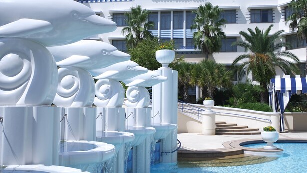 A pool area with multiple water features, including dolphin sculptures and fountains, surrounded by palm trees and a hotel building with balconies. - Disney Ambassador Hotel (Urayasu)