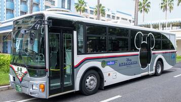 Un autobus di grandi dimensioni con la scritta Disney Ambassador Hotel, parcheggiato in strada con palme e un edificio alberghiero sullo sfondo.