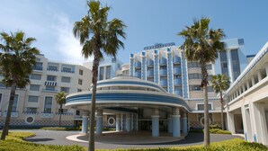 A hotel with a curved entrance, palm trees, and a multi-story building with a blue and white facade. - Disney Ambassador Hotel (Urayasu)