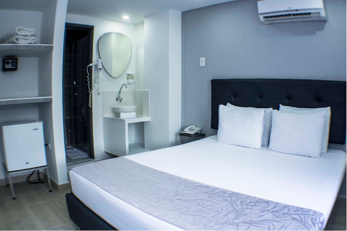 Habitación de hotel con una cama grande, teléfono junto a la cama, espejo, mueble blanco pequeño, lavamanos y unidad de aire acondicionado.
