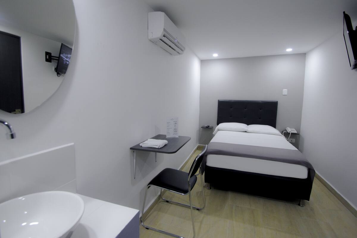 Una habitación de hotel moderna con cama, mesita de noche, silla y baño con lavamanos y espejo.
