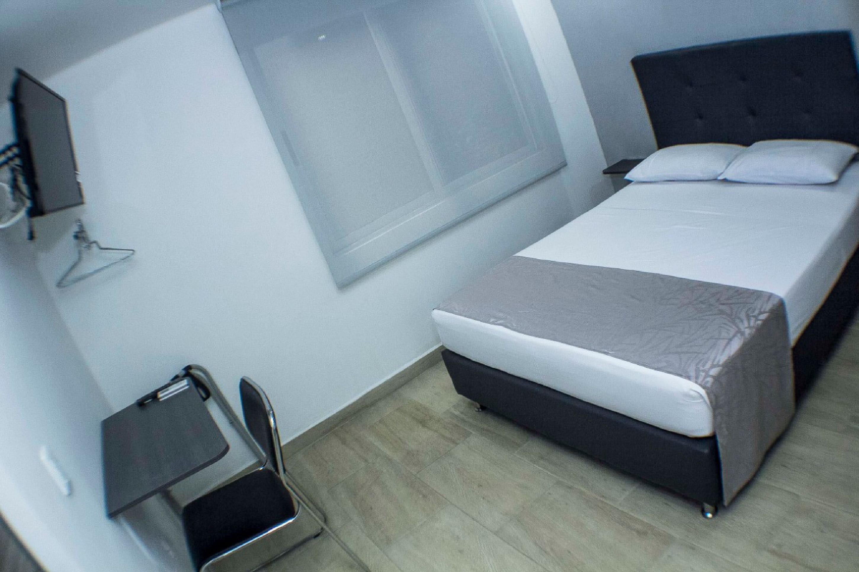 Una cama individual con cabecera gris, una mesita de noche pequeña, una silla y un televisor fijado a la pared.