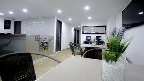 A hotel reception area with a reception desk, a seating area with chairs, a dining table, and a television mounted on the wall.