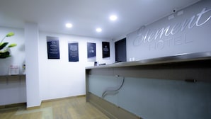 A hotel reception area with a reception desk, informational posters on the wall, and a wooden floor.