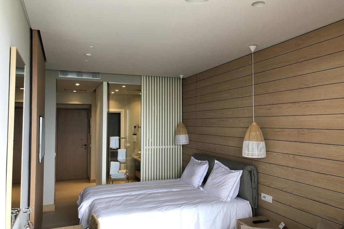 A modern hotel room with a large bed, wooden panel walls, a hanging light fixture, and a visible bathroom with a shower and towel rack.