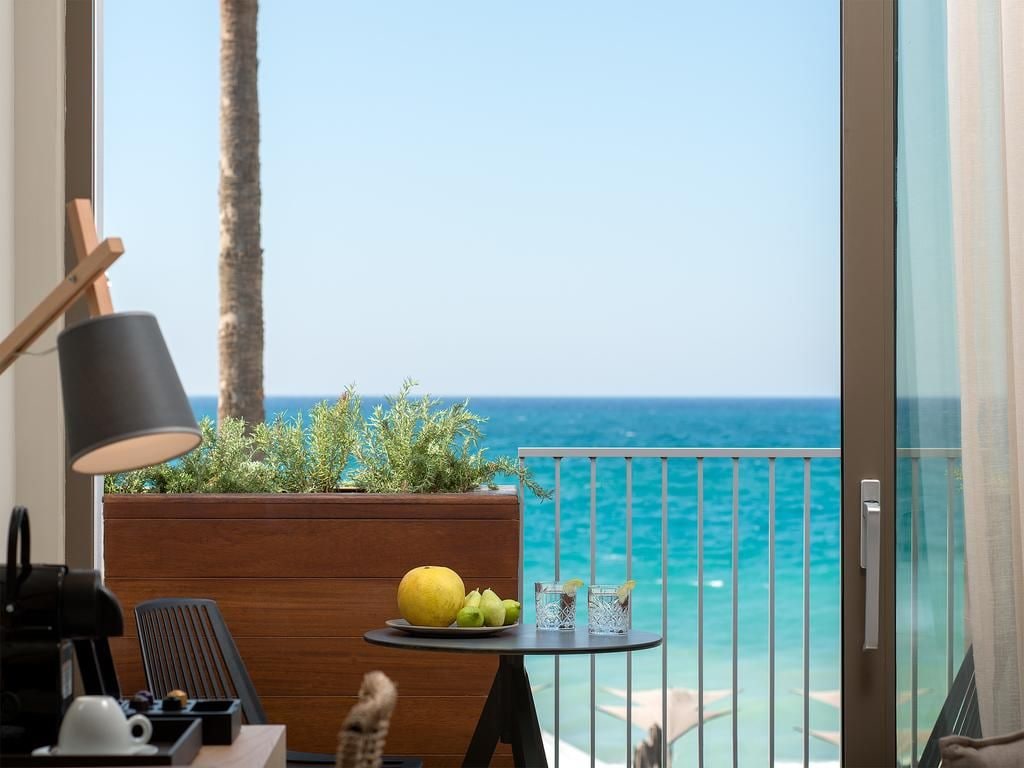 Un balcón con vistas al mar, una pequeña mesa con un frutero y vasos, una lámpara y una maceta de madera con plantas.