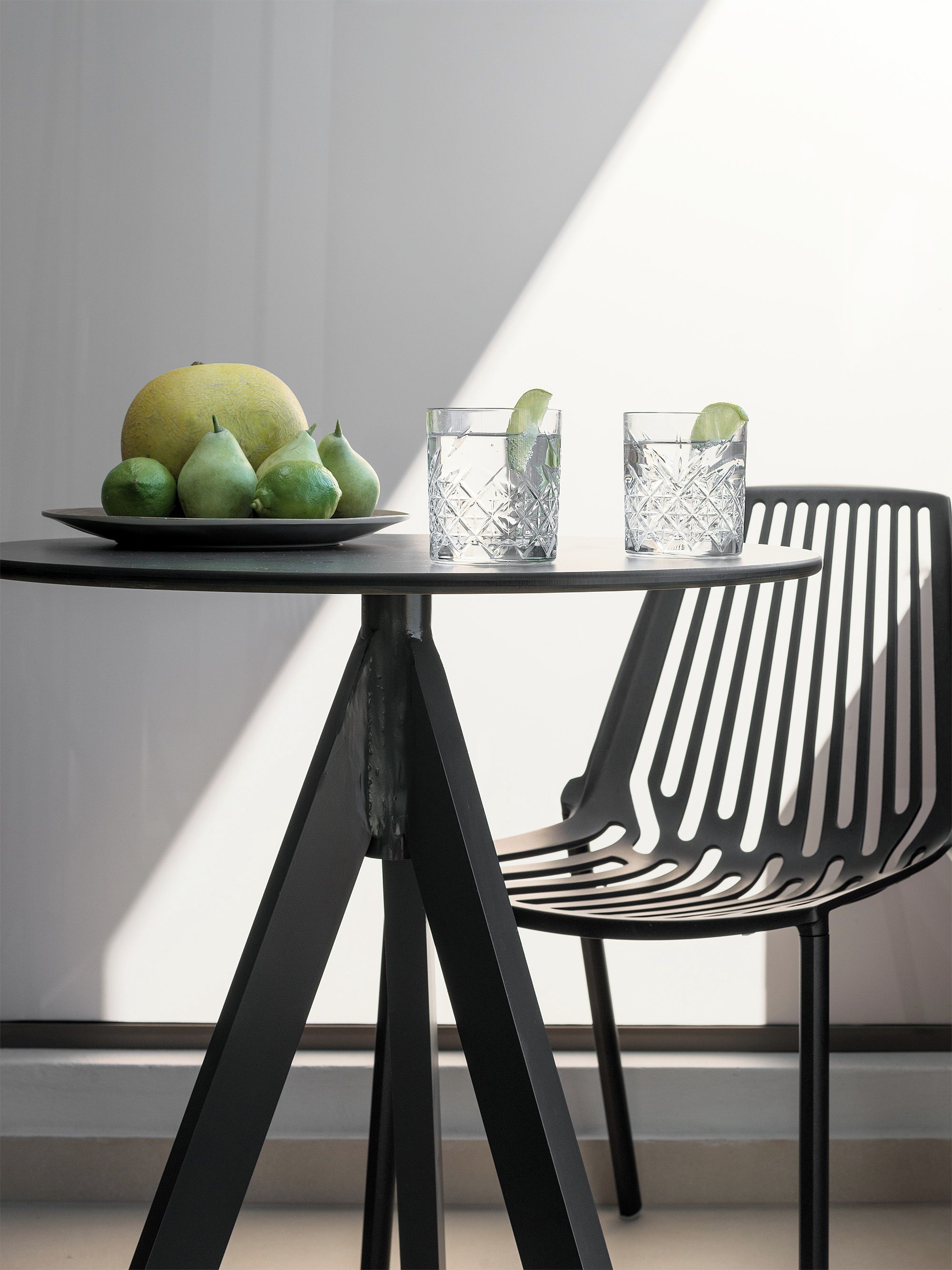 A table with a plate of fruit and two glasses of water, a black chair, and a window casting a shadow.