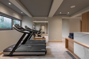 Une salle de sport avec des tapis de course alignés le long d’un couloir, un comptoir d’accueil avec un dessus en marbre et une fenêtre donnant sur un espace verdoyant.