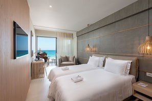 A modern hotel room with two beds, a TV, a balcony with a view of the sea, and a desk with a chair. - Ikones Seafront Luxury Suites (Rethymno)