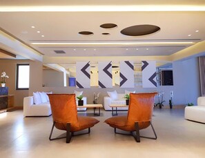 A modern hotel lobby with a seating area featuring two orange chairs, a white sofa, and a coffee table. - Ikones Seafront Luxury Suites (Rethymno)