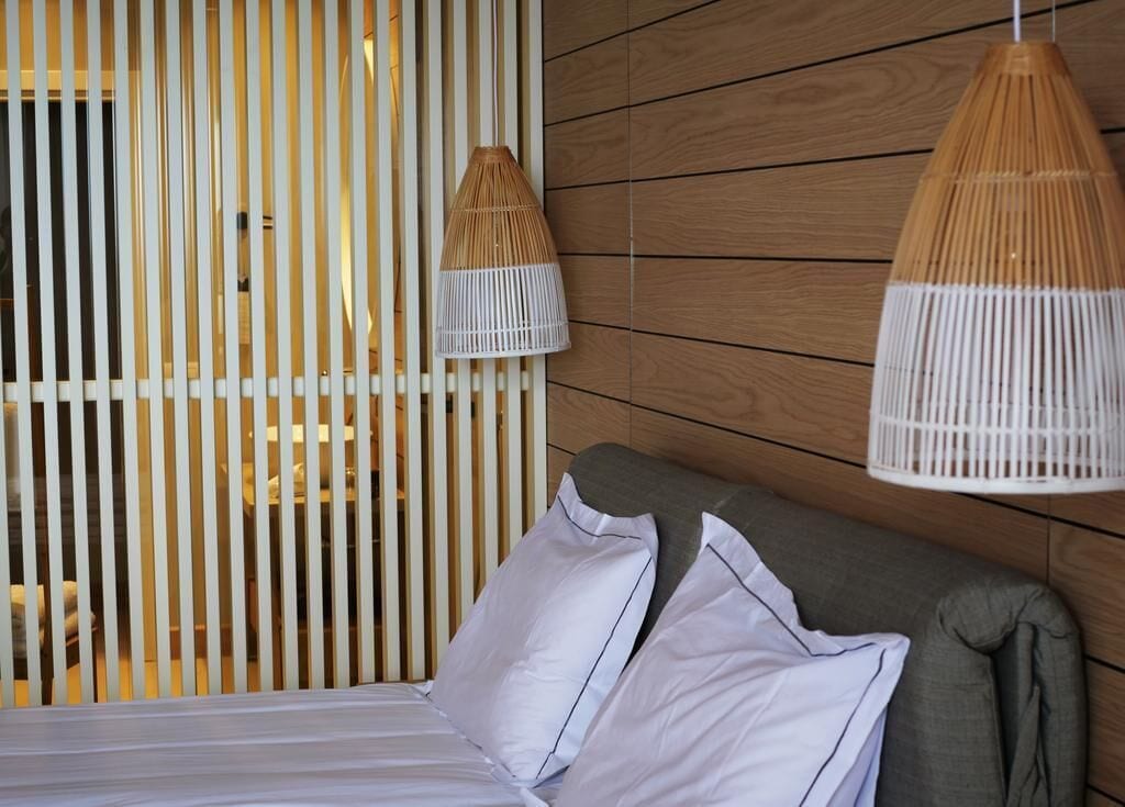 Una cama con sábanas blancas y dos almohadas, pared de madera y dos lámparas de mimbre colgantes.