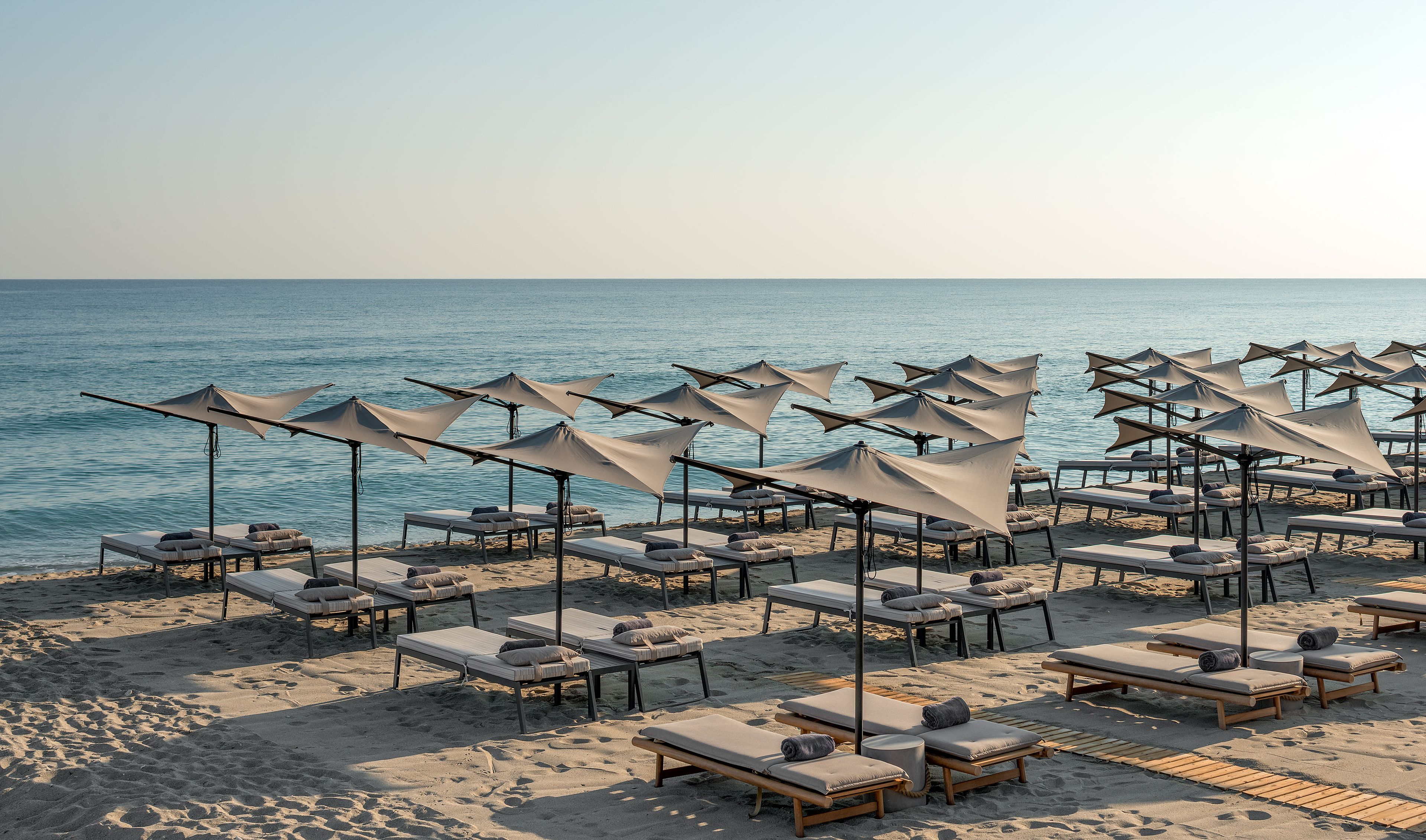 Ein Strand mit Reihen von Liegestühlen und Sonnenschirmen, einige mit Kissen, und einem ruhigen Meer im Hintergrund.
