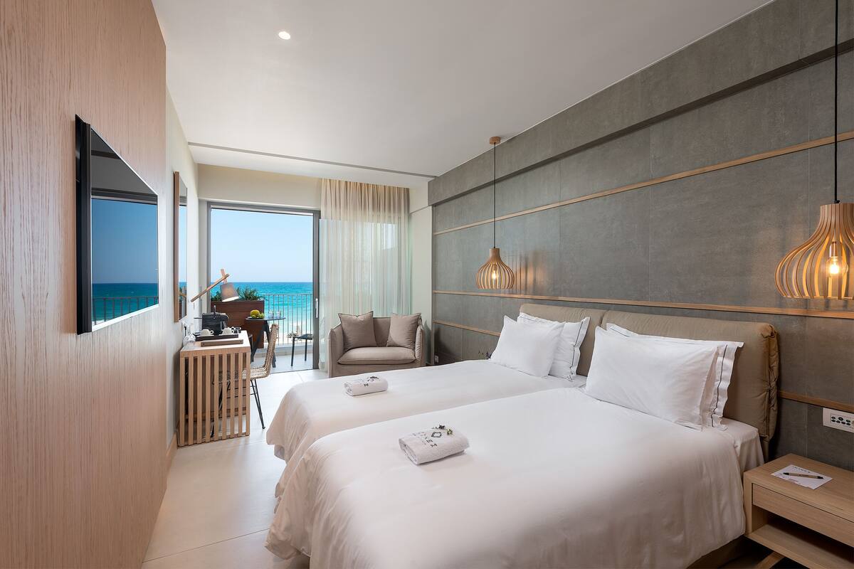 Een moderne hotelkamer met twee bedden, een televisie, een balkon met uitzicht op zee, en een bureau met stoel.