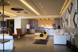 A modern hotel lobby with a reception desk, sofas, a coffee table, and a waiter. - Ikones Seafront Luxury Suites (Rethymno)