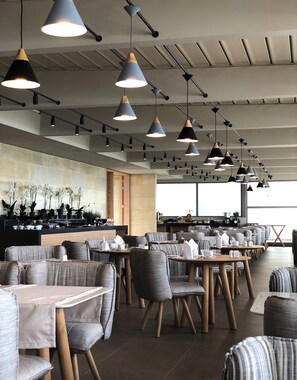 A modern restaurant interior with multiple dining tables and chairs, pendant lighting, and a counter with plants. - Ikones Seafront Luxury Suites (Rethymno)