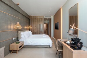 A modern hotel room with a bed, bedside table, desk, chair, and a large mirror. - Ikones Seafront Luxury Suites (Rethymno)