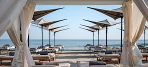 Un espace de détente aménagé sur la plage, avec des rangées de chaises longues à l’ombre de grands parasols, offrant une vue sur la mer.