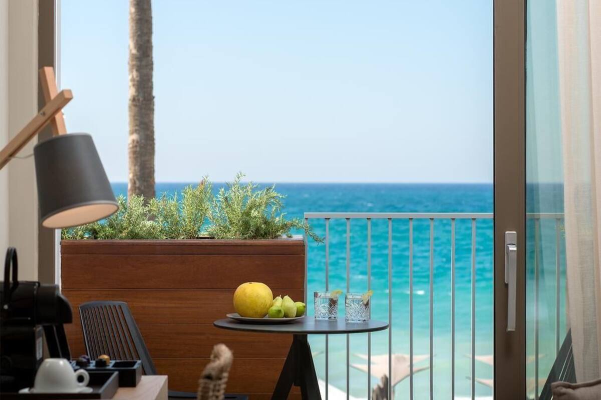 Een balkon met uitzicht op zee, een kleine tafel met een schaal fruit en glazen, een lamp, en een houten plantenbak met planten.