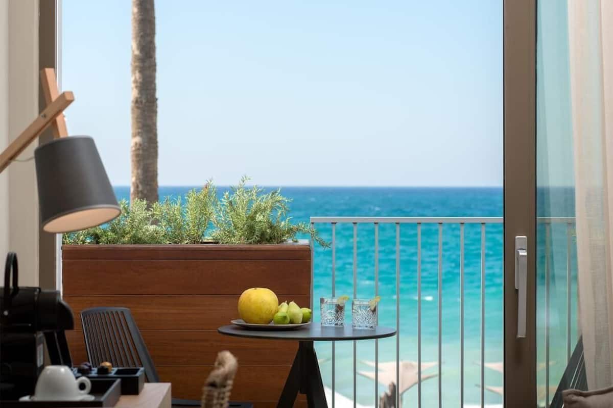 A balcony with a view of the sea, a small table with a fruit bowl and glasses, a lamp, and a wooden planter with plants.