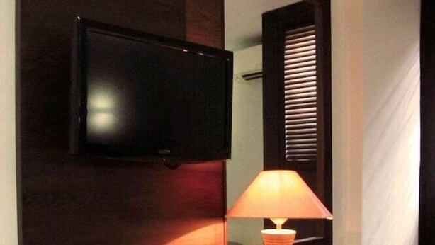 A hotel room with a dark wood desk, a lamp, a mirror, and a flat-screen TV. - Alma Hotel (Jakarta)