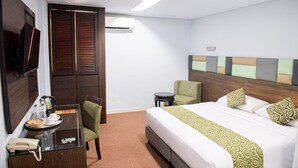 A hotel room with a bed, a desk with a telephone, a chair, a small table with a kettle and cups, and a television on the wall. - Alma Hotel (Jakarta)