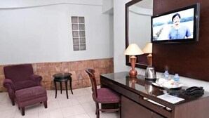 A hotel room with a desk, a chair, a small table, a lamp, and a television mounted on the wall. - Alma Hotel (Jakarta)
