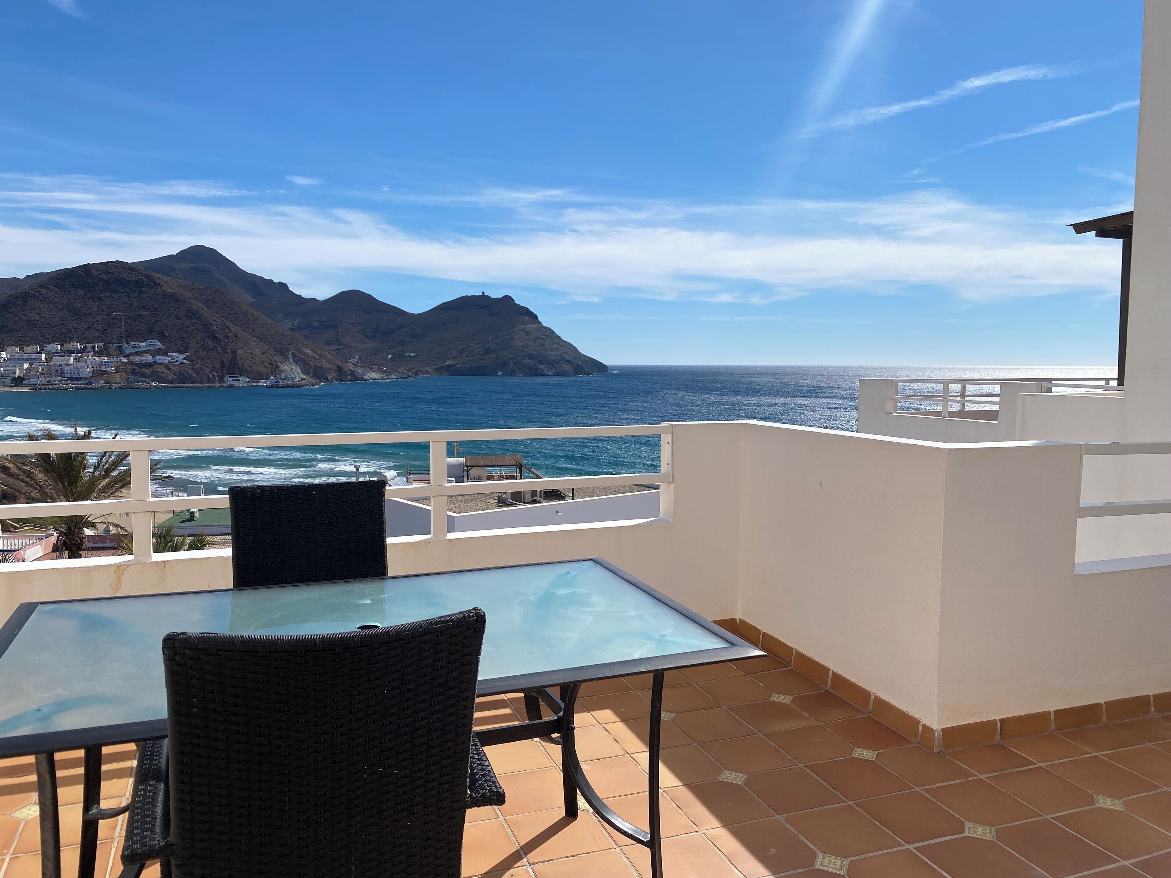 Een balkon met een glazen tafel en rieten stoel, met uitzicht op de kust, bergen en de zee.