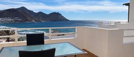 Un balcón con mesa de vidrio y silla de mimbre, con vistas a la costa, montañas y el mar.
