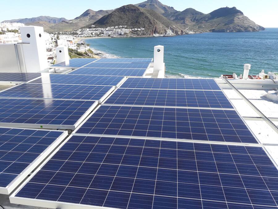 Paneles solares en un tejado con un pueblo costero y montañas al fondo.