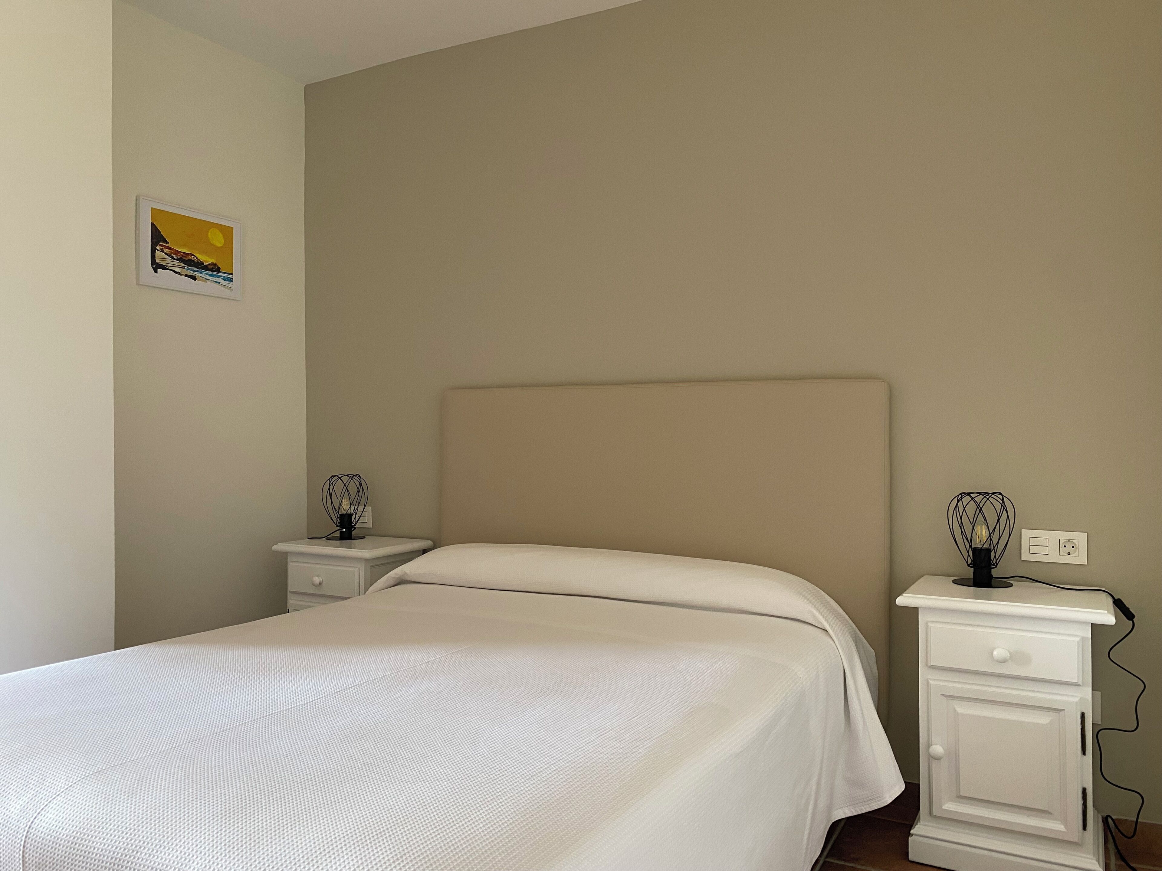 Un dormitorio con una cama, dos mesitas de noche y una mesita auxiliar con una lámpara.