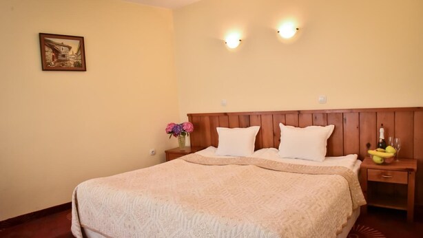 A neatly made bed with a wooden headboard, a nightstand with a vase of flowers, and a small table with a bottle and glasses. - Lina Guest House (Bansko)