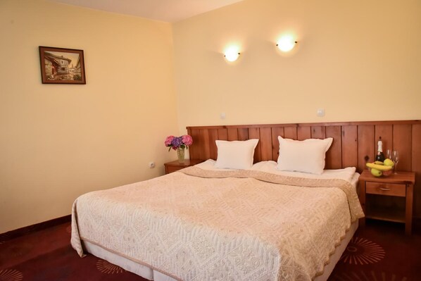 Cama bem arrumada com cabeceira de madeira, mesa de cabeceira com um vaso de flores e uma mesinha com garrafa e copos.