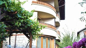 A multi-story building with a curved balcony, surrounded by lush greenery and flowering plants. - Lina Guest House (Bansko)