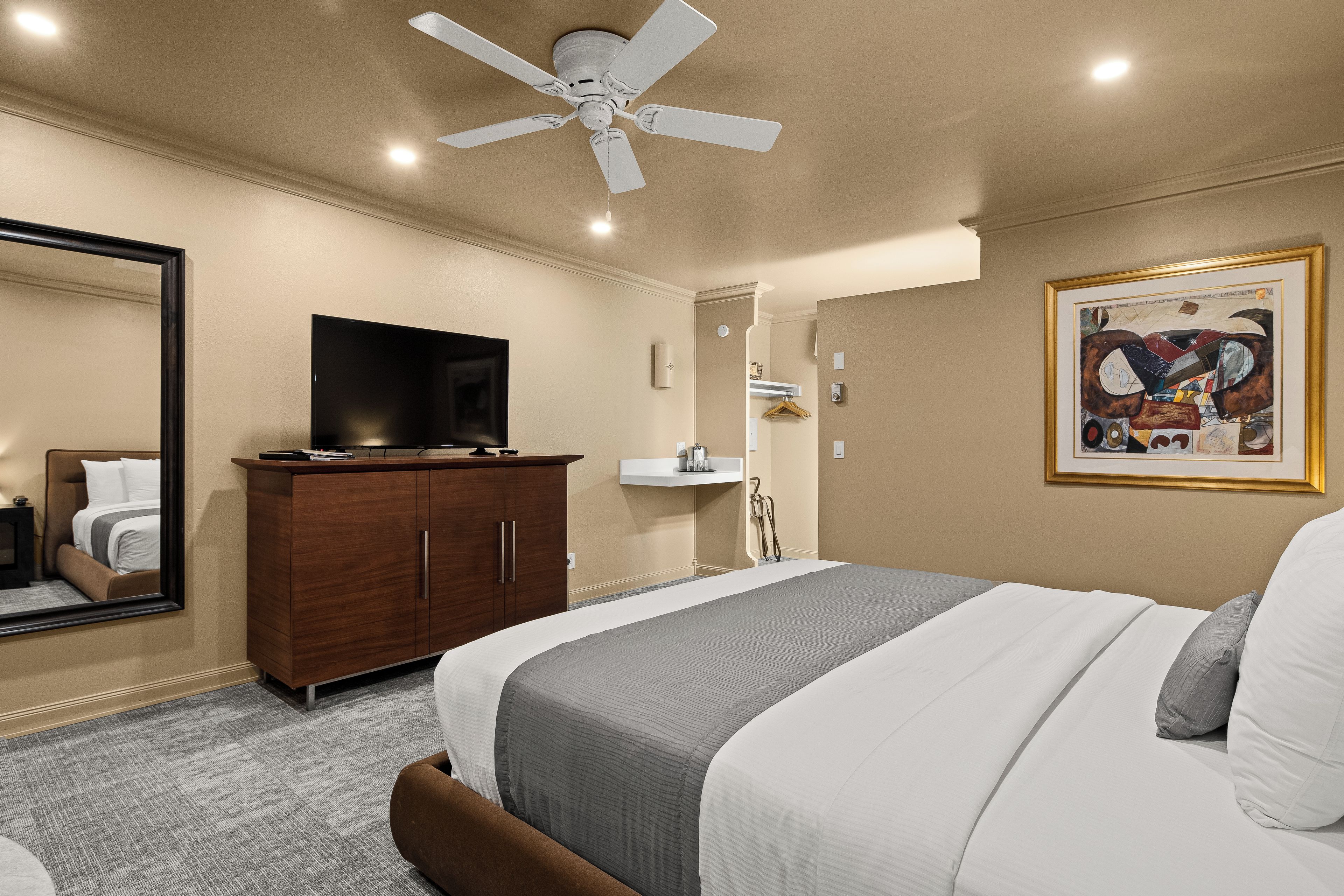 A hotel room with a bed, a television on a wooden cabinet, a ceiling fan, a bathroom with a sink and towel rack, and a framed artwork on the wall.