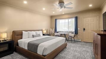 A hotel room with a bed, bedside tables, a desk, a chair, a window with curtains, a door, and a ceiling fan.