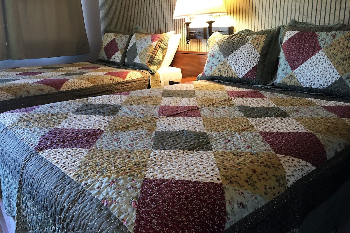 A hotel room with two beds, each covered with a quilt featuring a patchwork design. There is a nightstand with a lamp between the beds.