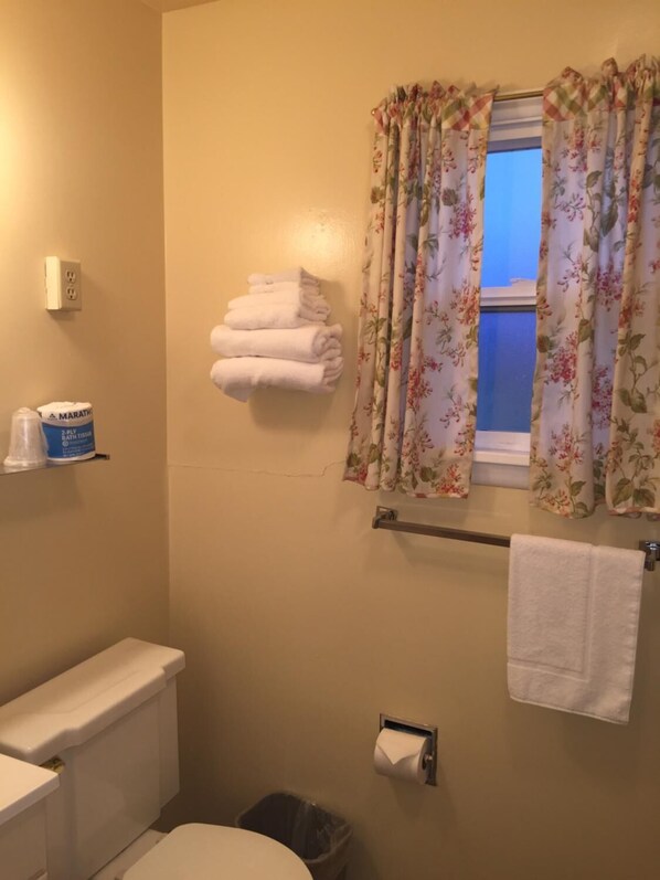 A bathroom with a toilet, a trash can, a towel rack with folded towels, a window with floral curtains, and a shelf with a container.