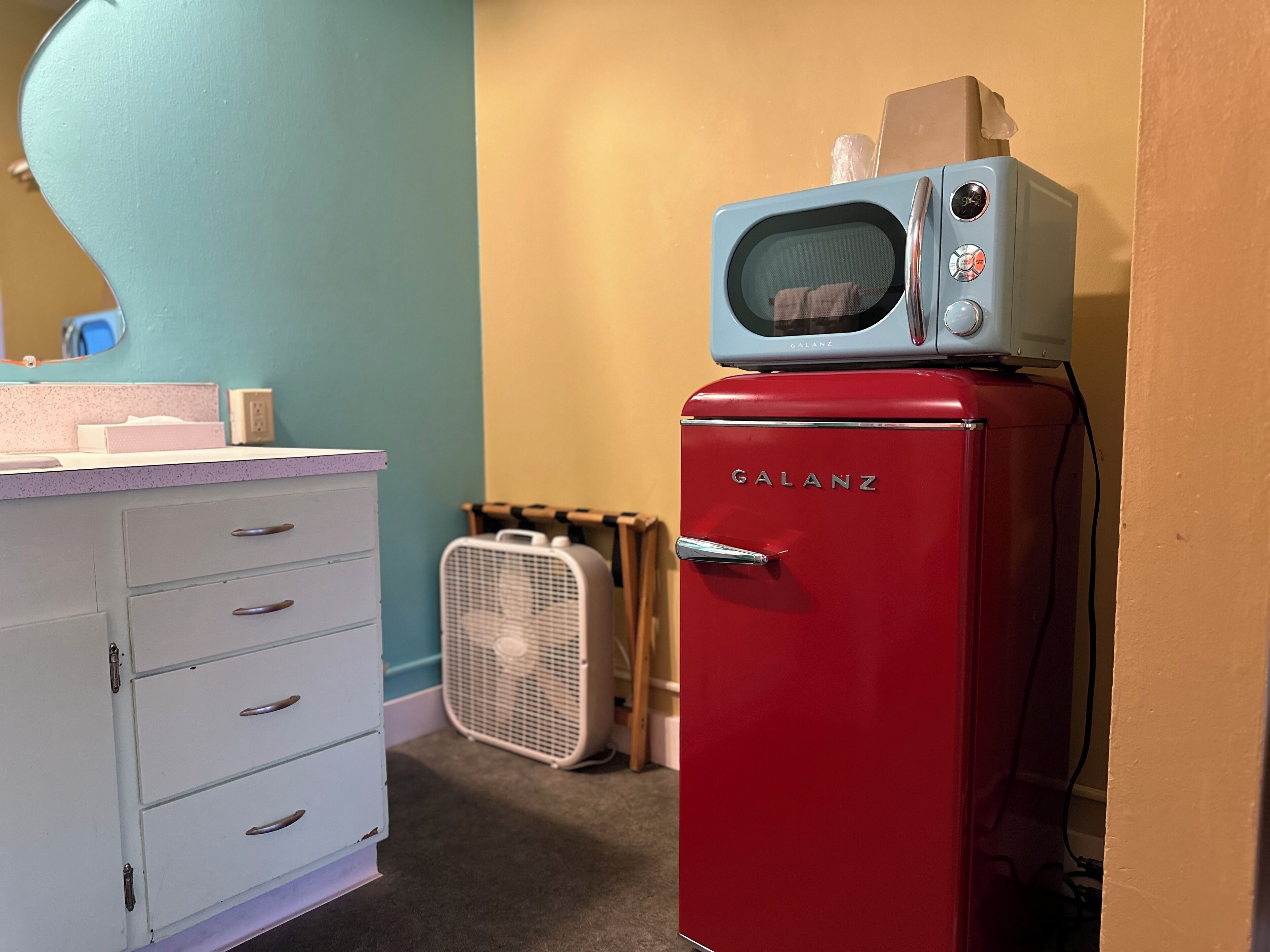 A red Galanz refrigerator with a microwave on top, a white fan, and a light blue cabinet with drawers.