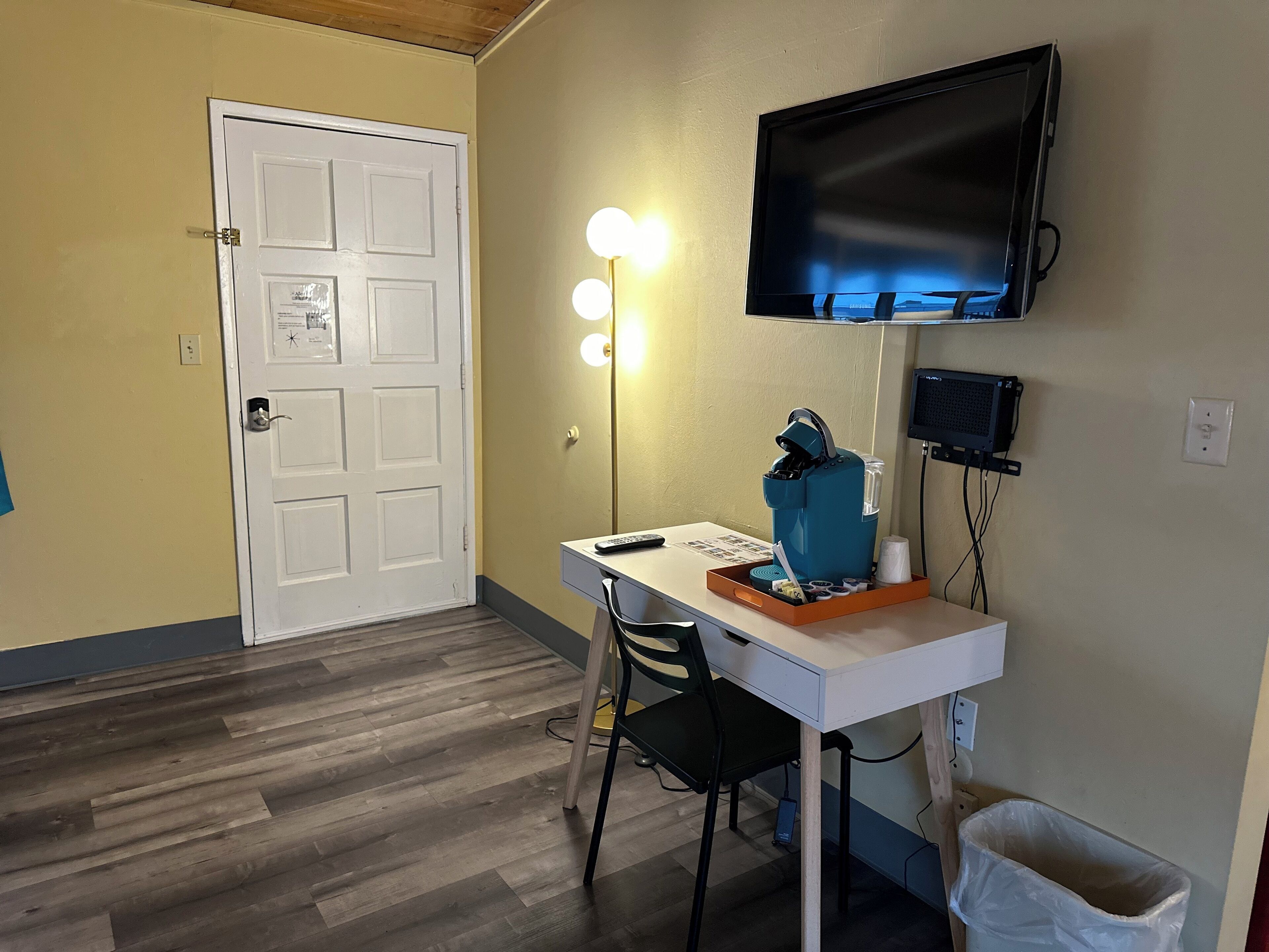 Um quarto pequeno com uma escrivaninha branca, uma cadeira, uma TV fixada na parede, uma cafeteira e um cesto de lixo.