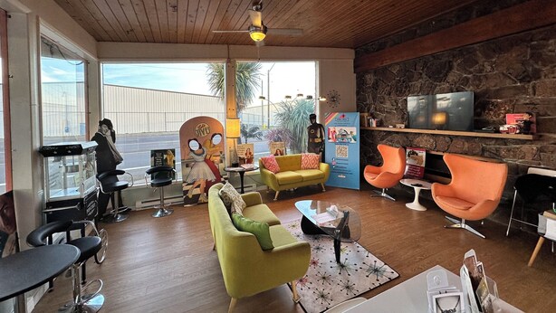 A spacious living area with a wooden ceiling, a stone wall, and a television mounted on the wall. There are two yellow sofas, a green sofa, and a black bar stool. A mannequin is visible near the entrance.