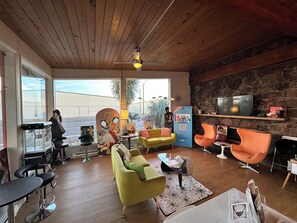 A spacious living area with a wooden ceiling, a stone wall, and a television mounted on the wall. There are two yellow sofas, a green sofa, and a black bar stool. A mannequin is visible near the entrance. - Atomic Motel (Astoria)