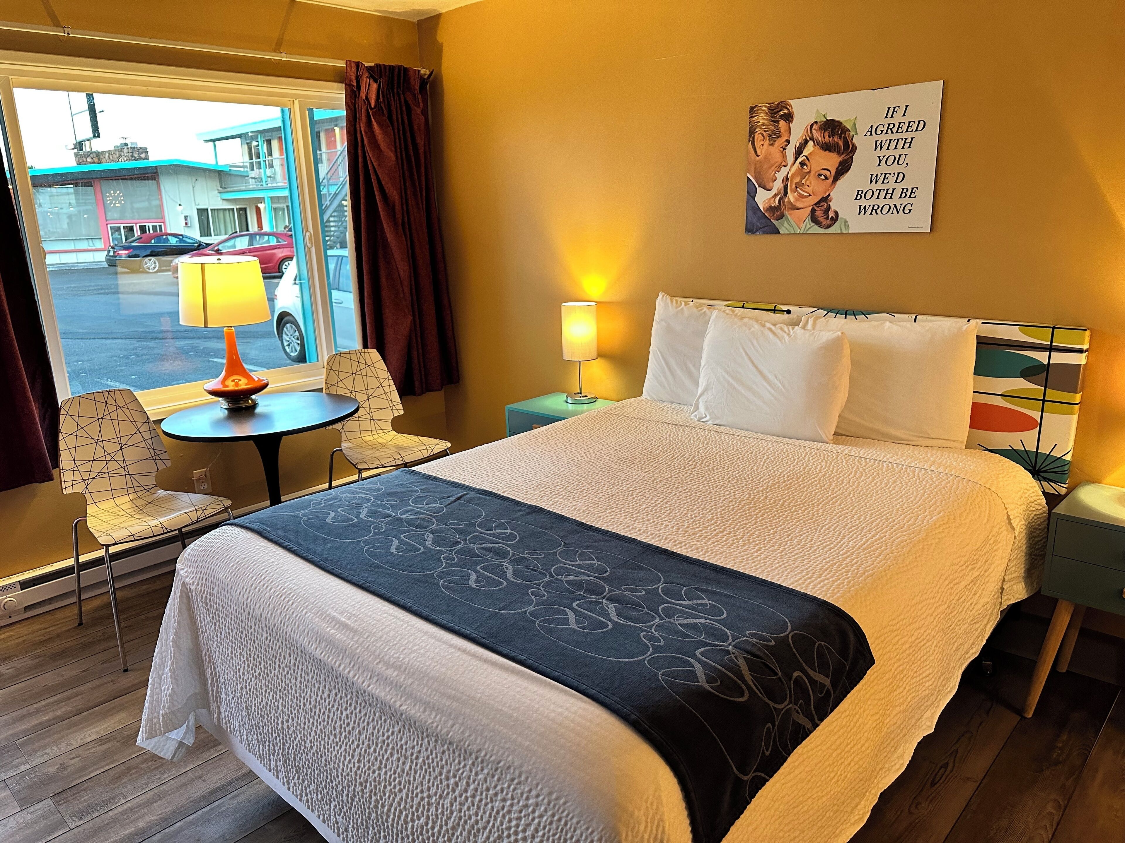 Habitación de hotel con una cama, una mesita con lámpara, dos sillas y vista a la calle.