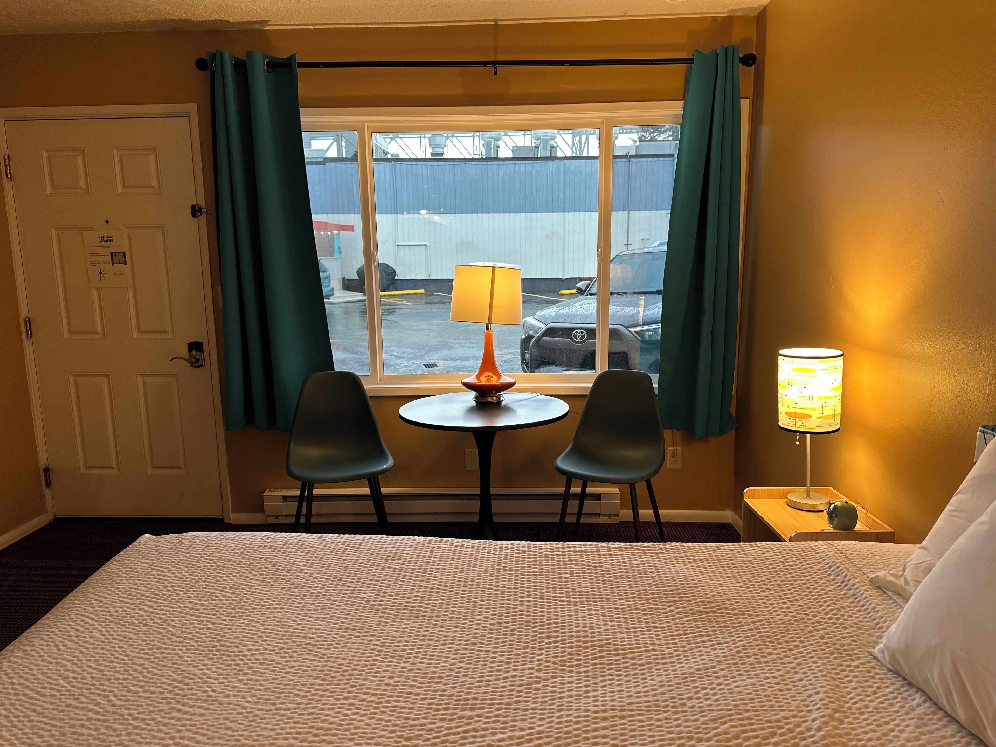 Een hotelkamer met een bed, twee stoelen, een tafeltje met een lamp en een raam met gordijnen.
