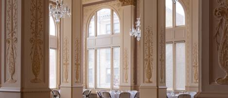 A spacious room with high ceilings, ornate columns, and large windows. Tables and chairs are arranged for dining.