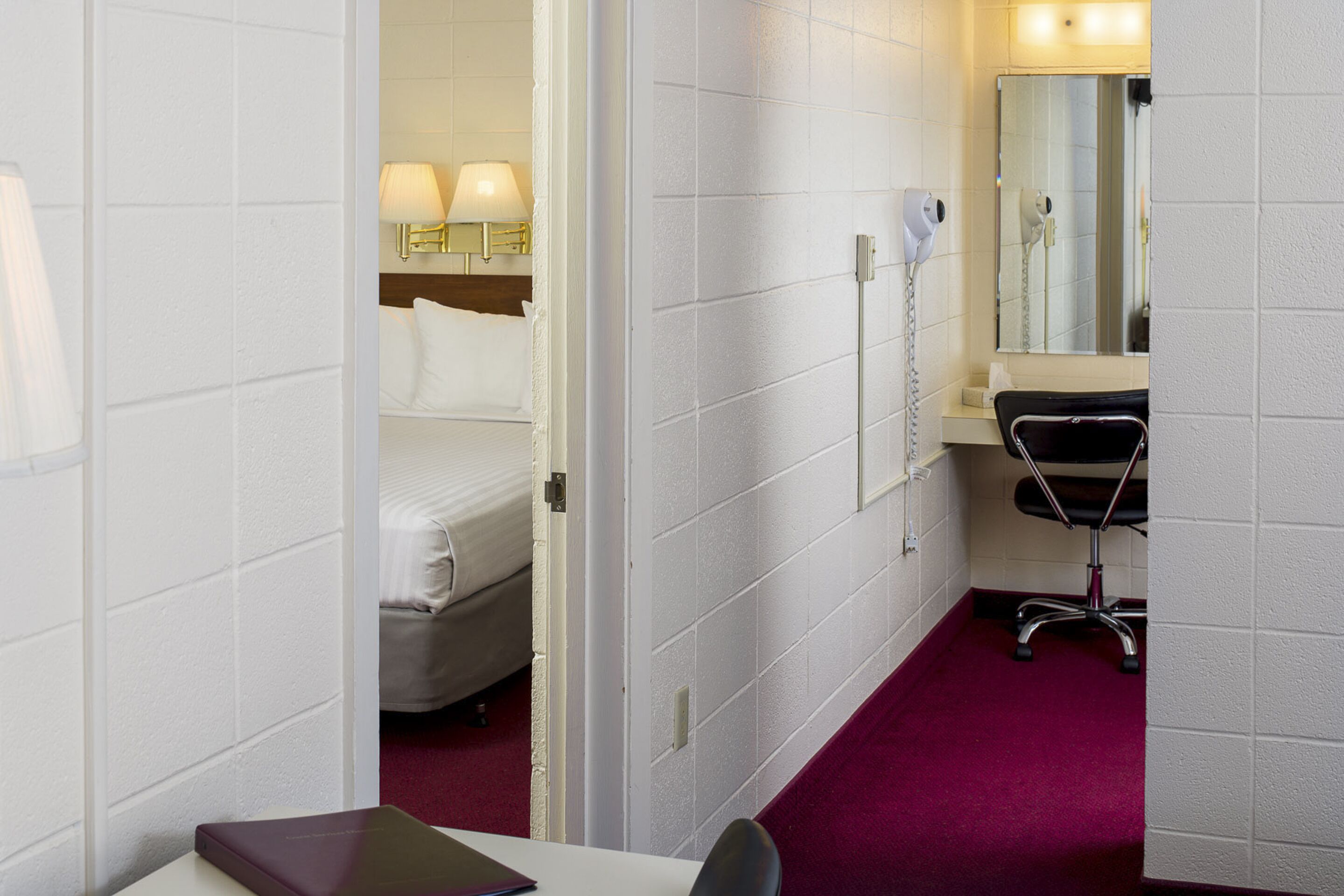 A hotel room with a bed, a desk with a chair, a desk lamp, a hairdryer, and a mirror.
