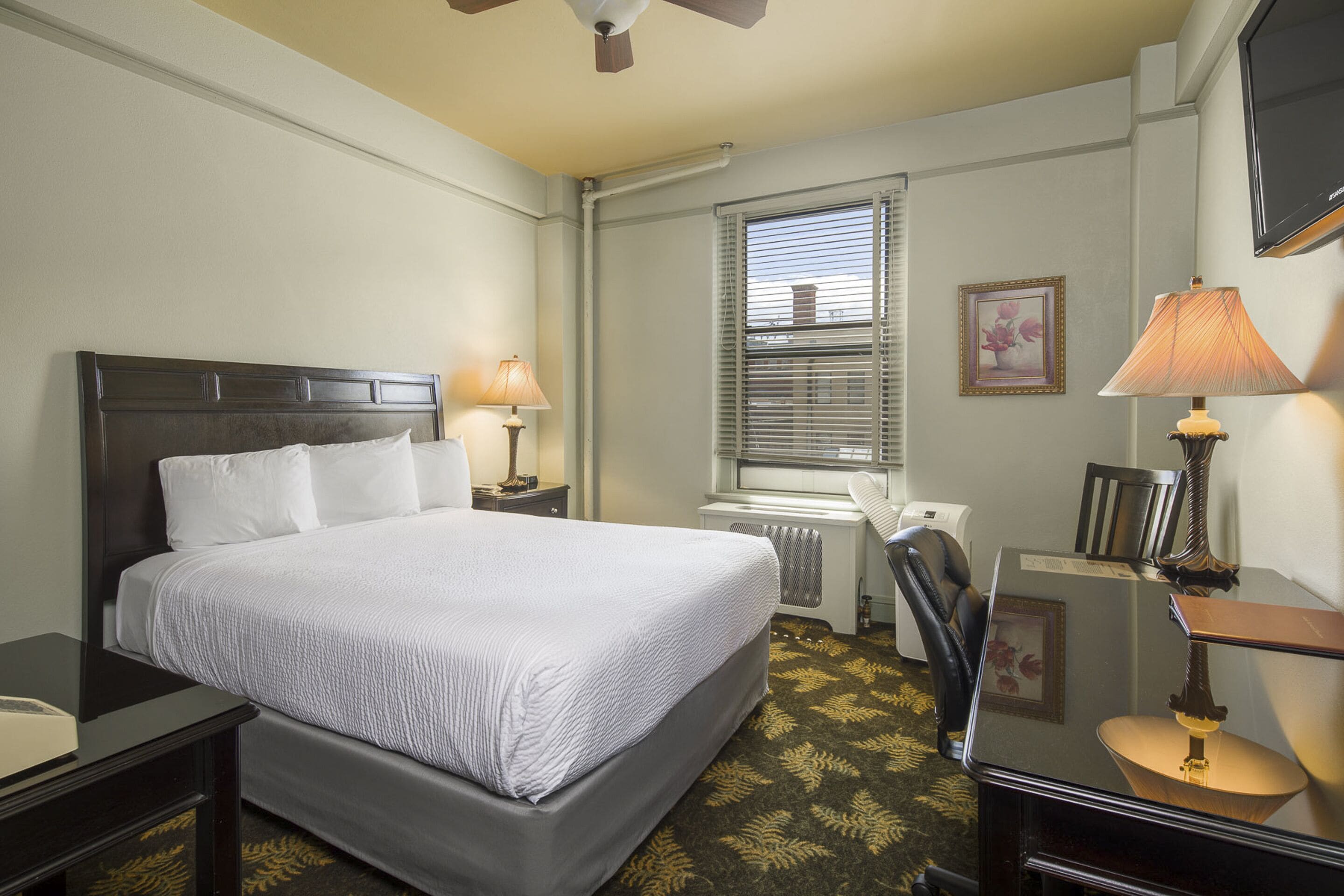 A hotel room with a large bed, a desk, a chair, a lamp, a radiator, a window with blinds, and a television on the wall.