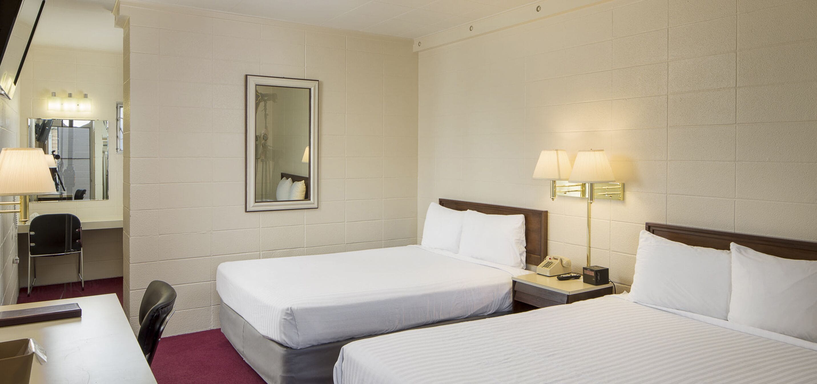 A hotel room with two single beds, a desk with a chair, a mirror, and a nightstand with a lamp and a phone.