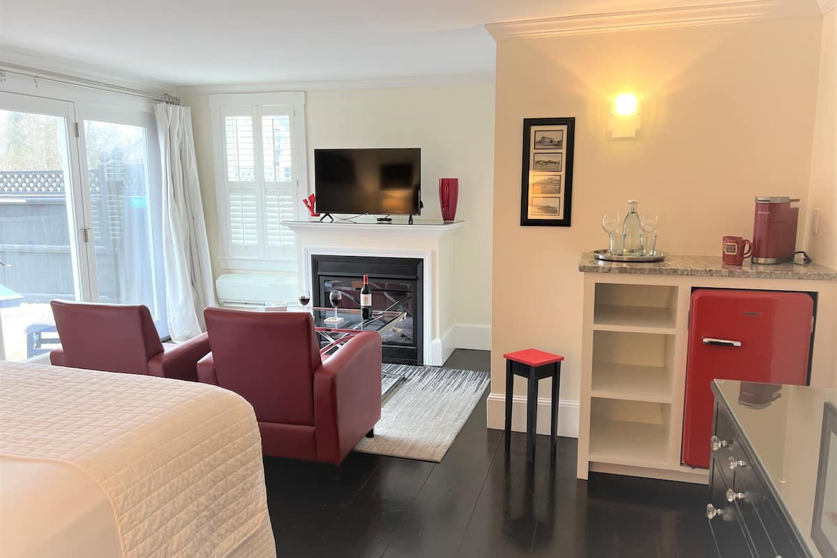 Habitación de hotel moderna con cama, zona de estar con televisor, chimenea, kitchenette y vistas al exterior.